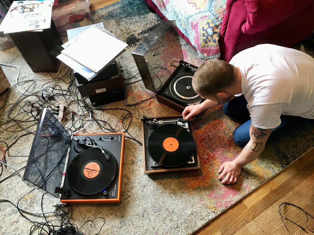 A man in a white t-shirt and jeans has three turntables and wires around him on the floor.